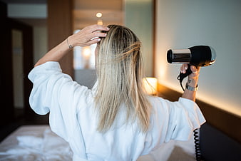 Woman in white robe blow-drying blonde hair in hotel room
