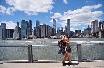 Two women hugging on a pier with the New York City skyline and Brooklyn Bridge