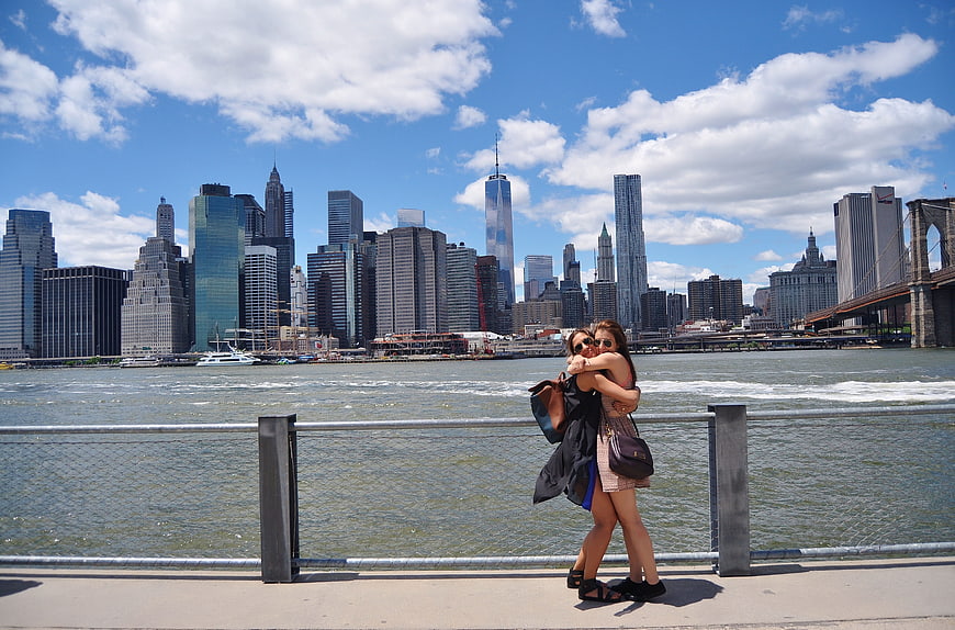 Two women hugging on a pier with the New York City skyline and Brooklyn Bridge