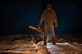 Scary hooded figure holding chainsaw in snowy night