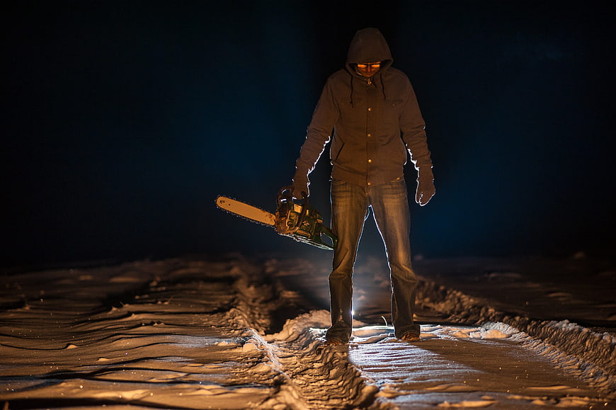Scary hooded figure holding chainsaw in snowy night