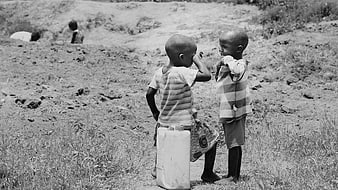 Two young African children standing in a rural field with a water container