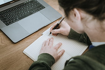 Over-the-shoulder view of person writing in notebook with laptop