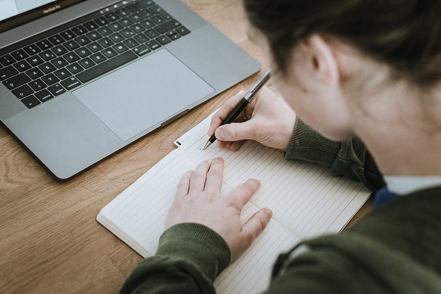Over-the-shoulder view of person writing in notebook with laptop