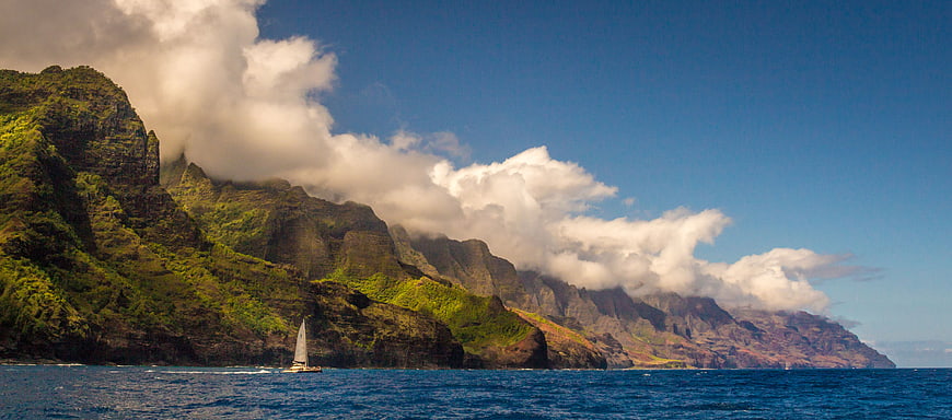 Sailboat sailing past dramatic green mountains and ocean