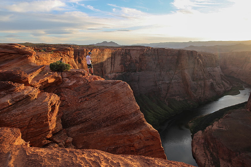 Hiker standing on red rock cliff overlooking river canyon at sunset