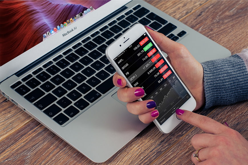 Woman checking stock market app on smartphone with laptop