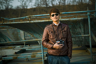 Man in Brown Leather Jacket Holding Camera on Rusty Bridge