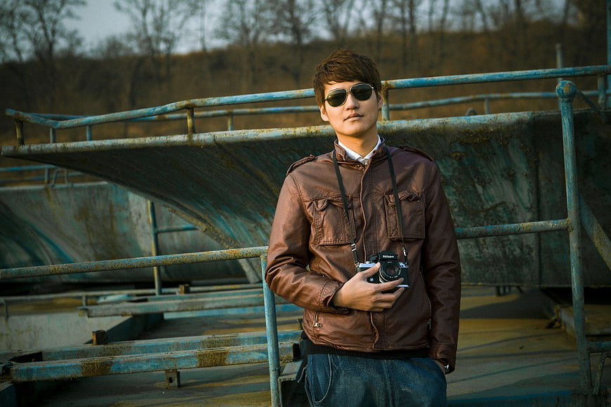 Man in Brown Leather Jacket Holding Camera on Rusty Bridge