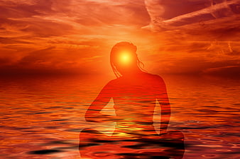 Silhouette of woman meditating in lotus position against fiery orange sunset ocean