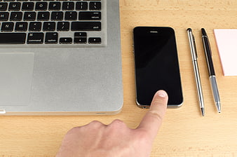 Hand pointing at smartphone screen on wooden desk with laptop
