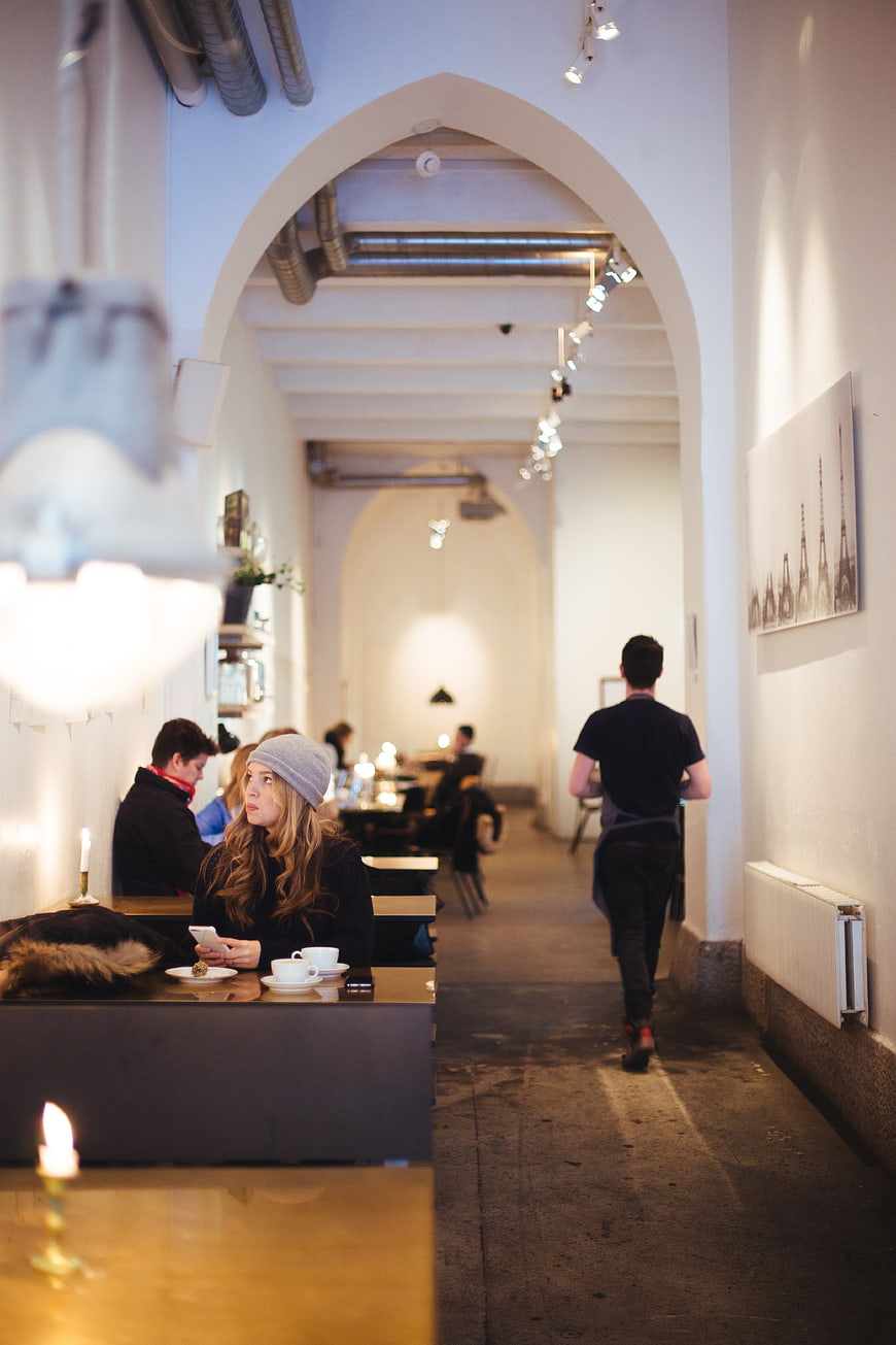 Woman in Beanie at Cafe Table with Waiter Walking Away