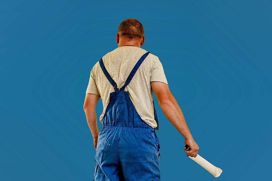 Handyman in blue overalls holding paint roller against blue wall