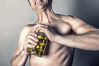 Shirtless muscular man holding a jar of pickles against a gray wall