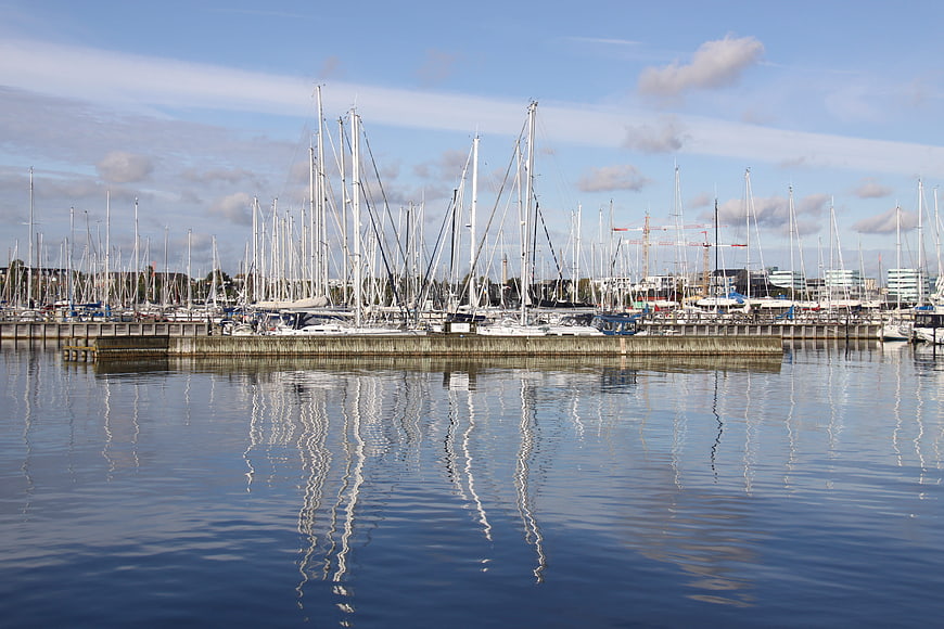 Sailboat masts reflecting in calm harbor water under blue sky