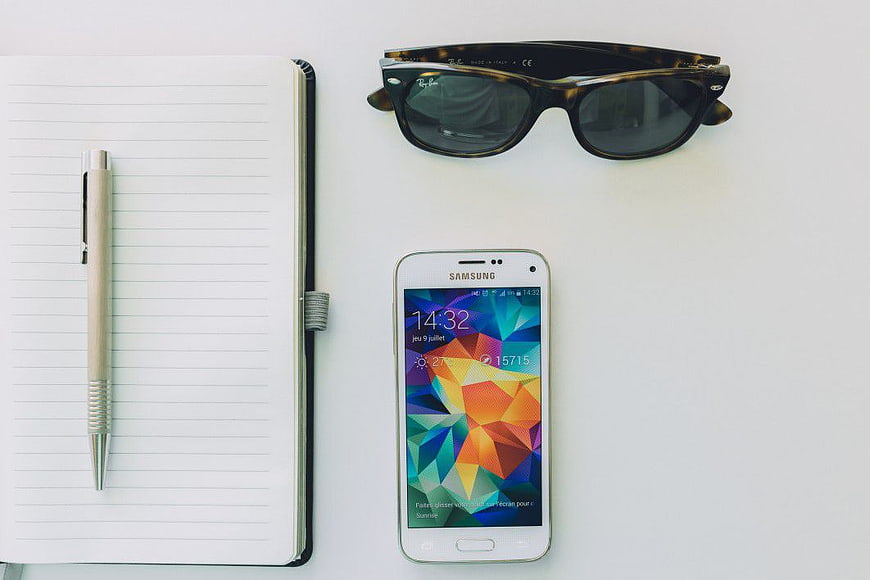 Flat lay of sunglasses, notebook, pen, and smartphone on white desk