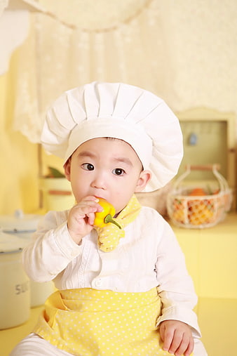 Cute baby chef eating yellow pepper in kitchen
