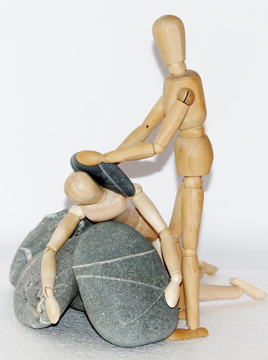 Wooden mannequin helping fallen figure with stones