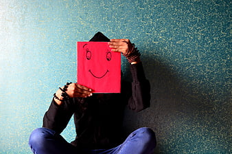 Person holding red smiley face paper mask against textured blue wall