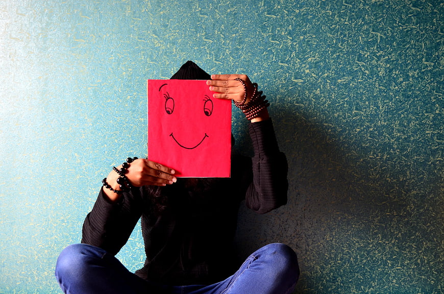 Person holding red smiley face paper mask against textured blue wall