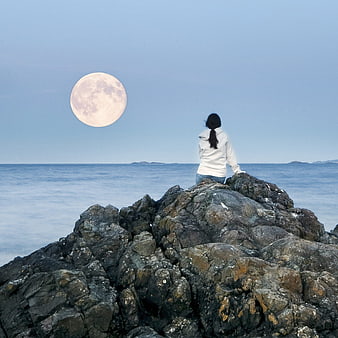 Woman sitting on rocks watching full moon over ocean
