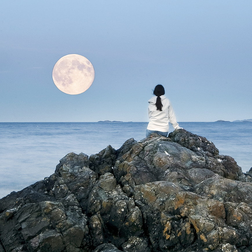 Woman sitting on rocks watching full moon over ocean