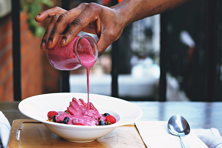 Hand Pouring Berry Sauce Over Fruit Dessert In White Bowl