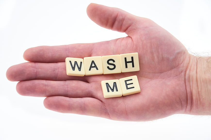 Hand holding Scrabble tiles spelling WASH ME on white background