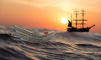 Paper boat on ocean waves with ship silhouette at sunset