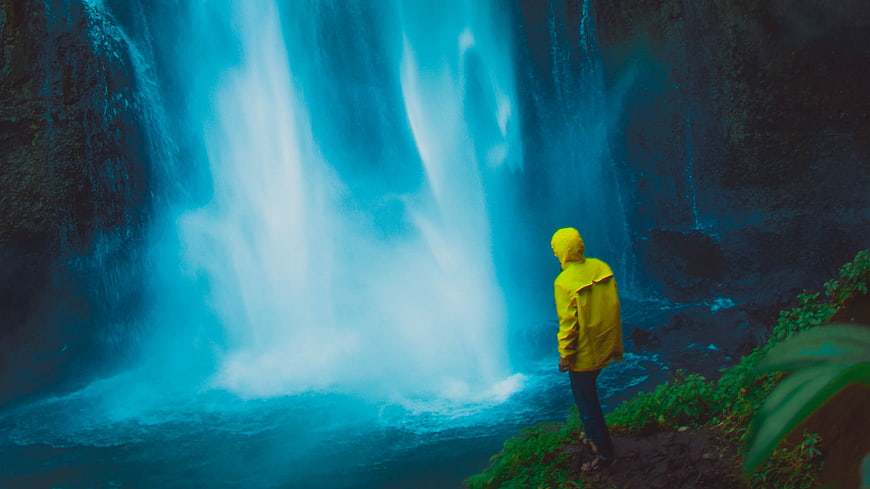 Man in Yellow Raincoat Standing Before Blue Waterfall