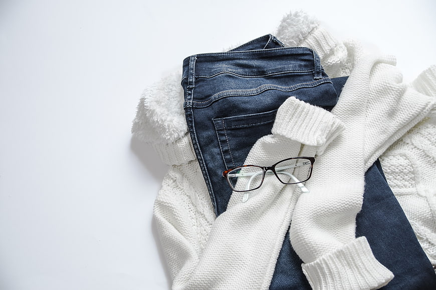 Flat lay of blue jeans, white sweater, and glasses on white background