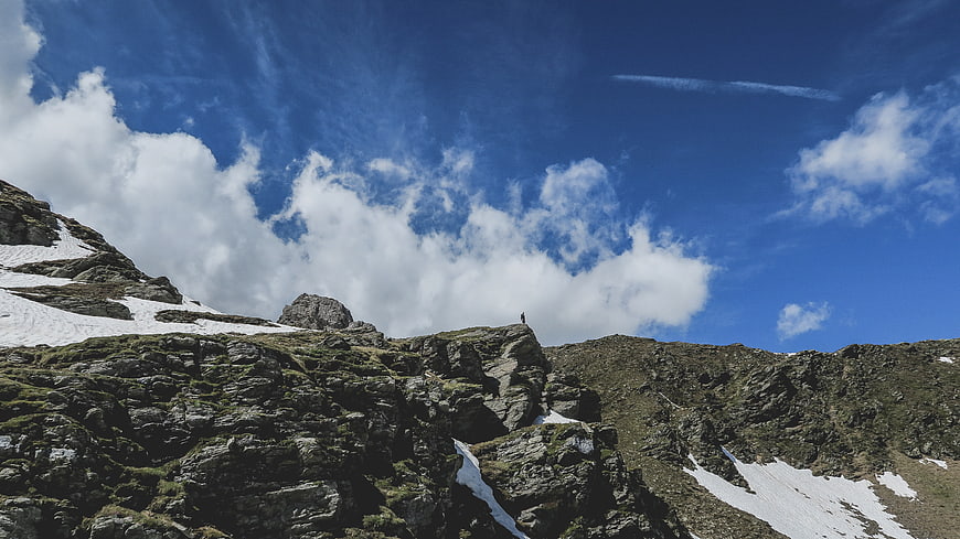 Lone Hiker Standing on Rocky Mountain Peak Under Blue Sky