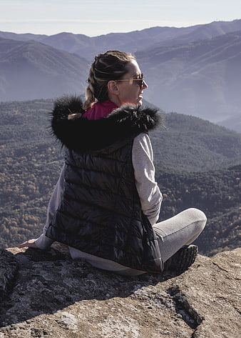 Woman in puffer vest sitting on mountain peak overlooking valley