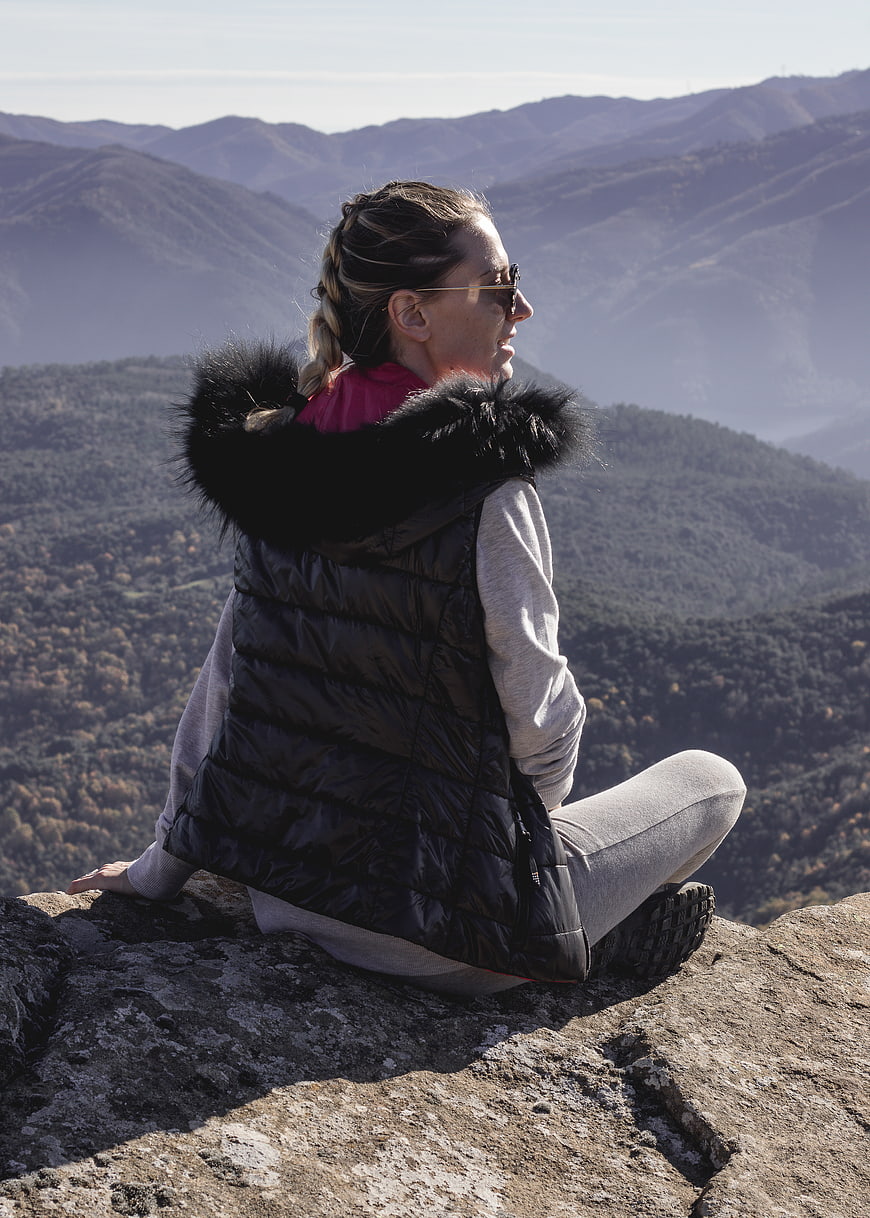 Woman in puffer vest sitting on mountain peak overlooking valley
