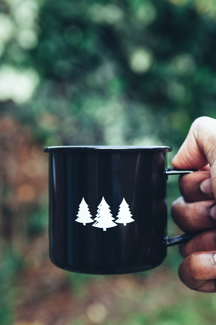 Hand holding black camping mug with white pine tree logo