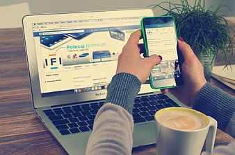 Person holding smartphone next to laptop and coffee on wooden desk