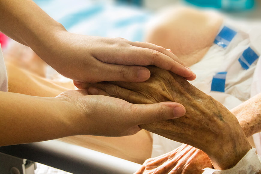 Young hands holding elderly patient hand in hospital bed