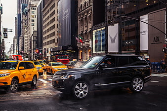 Black luxury SUV driving past yellow taxis on wet NYC street