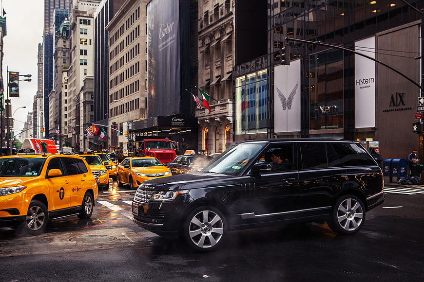 Black luxury SUV driving past yellow taxis on wet NYC street