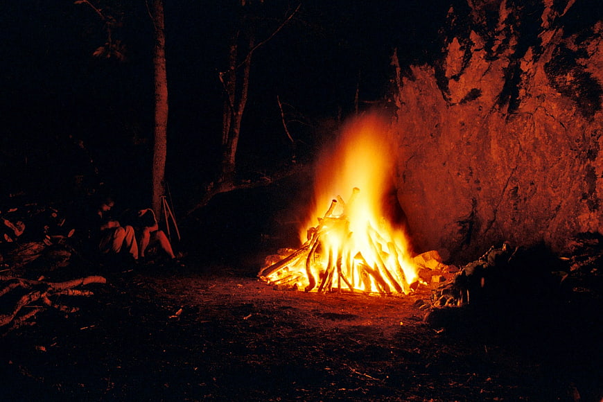 Large campfire burning at night in wilderness with people sitting nearby