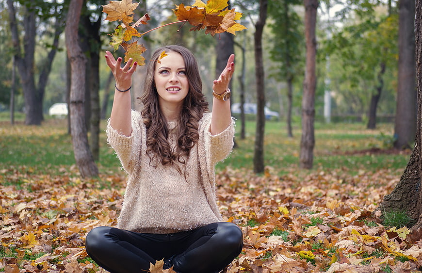 Young woman sitting in autumn park throwing maple leaves