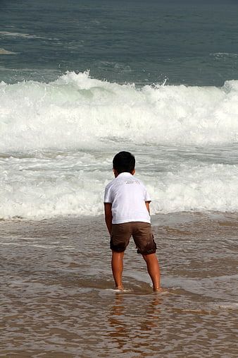 Child standing in shallow ocean water facing breaking waves