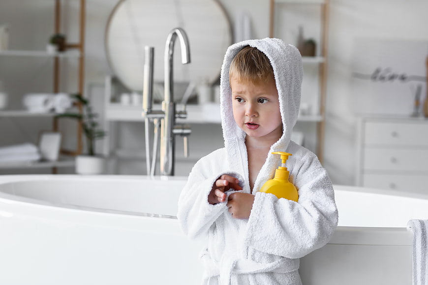 Toddler in white bathrobe holding yellow soap bottle in bathroom