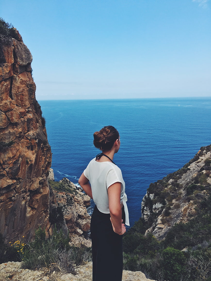 Woman in White Top Standing on Cliff Overlooking Blue Ocean