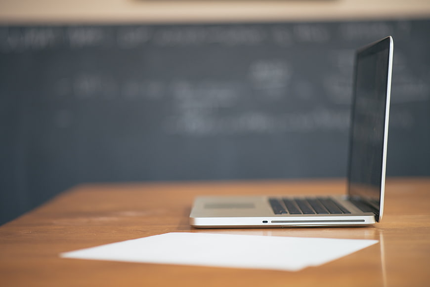 Open Silver Laptop On Wooden Desk With Blurred Chalkboard Background