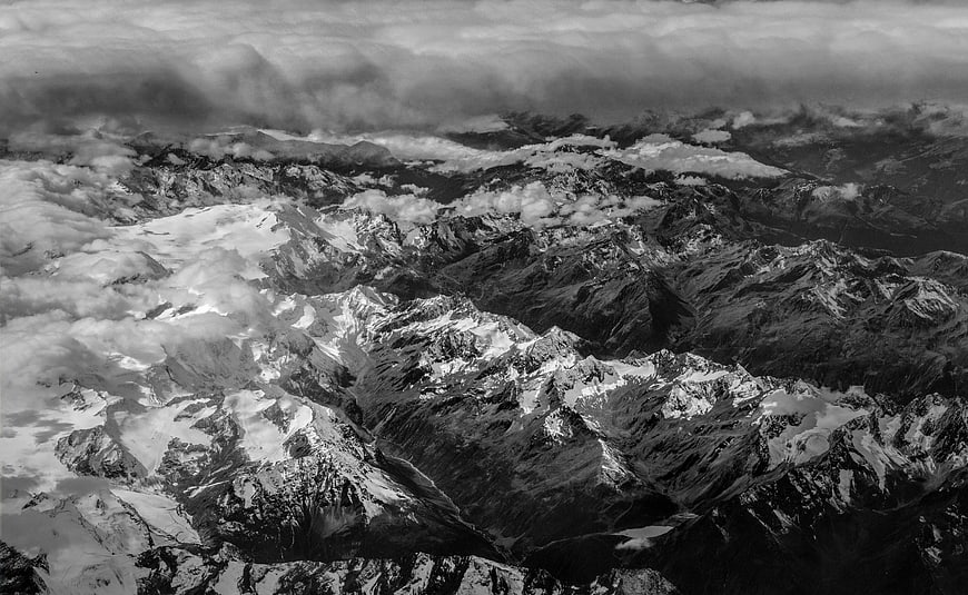 Black and white aerial view of snow-capped mountain peaks through clouds