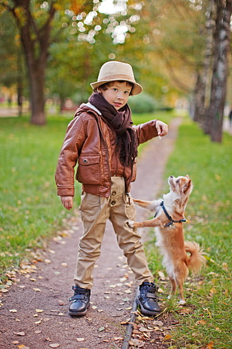 Young boy in leather jacket and hat playing with small dog in autumn park