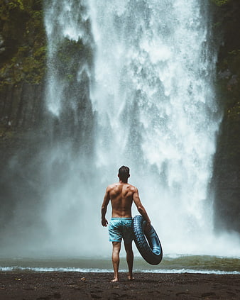 Man with inner tube standing before massive waterfall