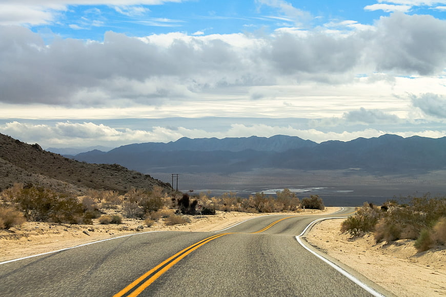 Scenic Desert Highway Leading To Distant Mountain Range Under Cloudy Sky
