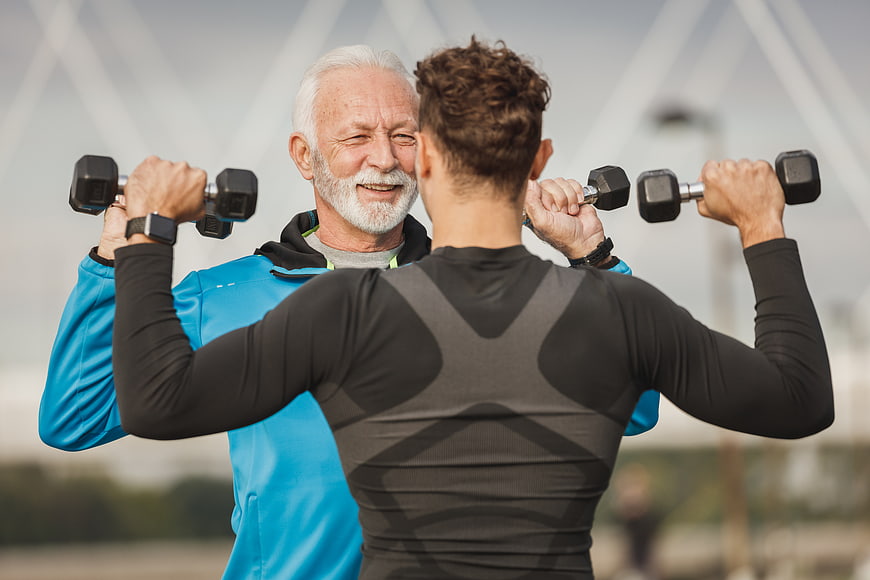 Senior man and young trainer lifting dumbbells outdoors